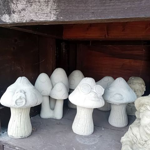 A group of unpainted grey concrete mushroom ornaments in several shapes and sizes on a wooden shelf, one with a face and glasses