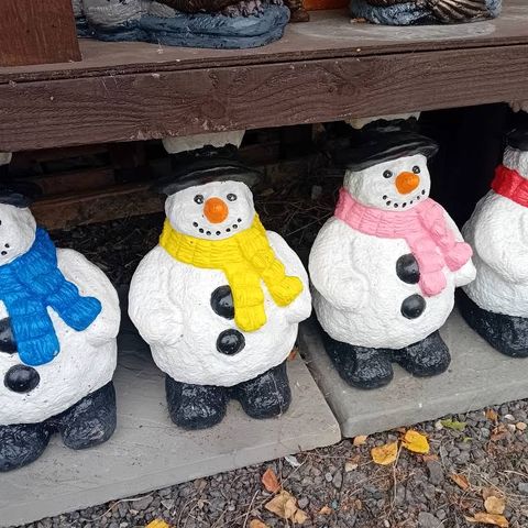 A row of hand-painted concrete snowmen with black top hats and blue, yellow and pink scarves standing on paving slabs under a wooden shelf