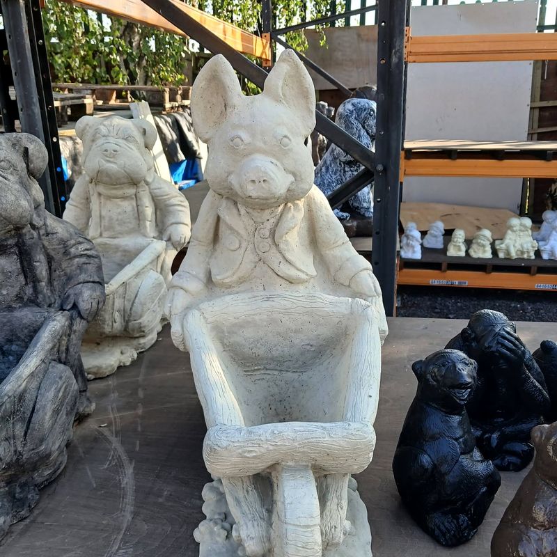 Two wheelbarrow planter figures in natural pale stone, with black monkey figures beside them and metal racking with shelves of ornaments behind
