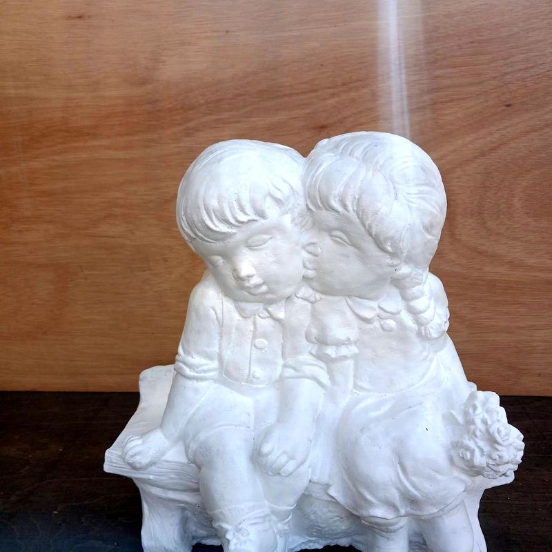 White concrete statue of a boy kissing a girl on the cheek as they sit on a bench, the girl holding a posy